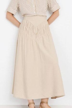 Mujer vistiendo falda midi beige con canesú en pico y detalle de frunces delanteros.